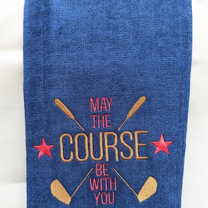 May include: A blue golf towel with the embroidered text "May the course be with you" in gold and red. Two red stars and two gold golf clubs are also embroidered on the towel.