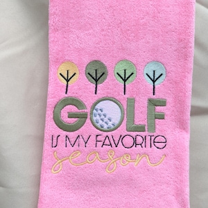 May include: Pink golf towel with embroidered text and graphics. The towel features the word "GOLF" with a golf ball graphic, and the phrase "IS MY FAVORITE SEASON". Above the text are embroidered trees.