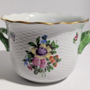 May include: A white porcelain bowl with a textured basketweave surface, featuring a central hand-painted floral bouquet in yellow, purple, pink, and green. The bowl has two green handles and a scalloped rim accented with a thin gold-tone trim.