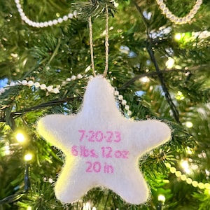 K&ouml;nnte beinhalten: Wei&szlig;er Filzstern-Anh&auml;nger mit rosa Stickerei. Der Anh&auml;nger zeigt "7.20.23 6 lbs, 12 oz 20 in".