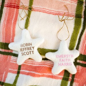 K&ouml;nnte beinhalten: Zwei wei&szlig;e sternf&ouml;rmige Ornamente mit goldener Schnur. Das Ornament links tr&auml;gt die Namen "Robin Jeffrey Scott" in goldenen Buchstaben. Das Ornament rechts tr&auml;gt die Namen "Emersyn Faith Harris" in rosa Buchstaben.