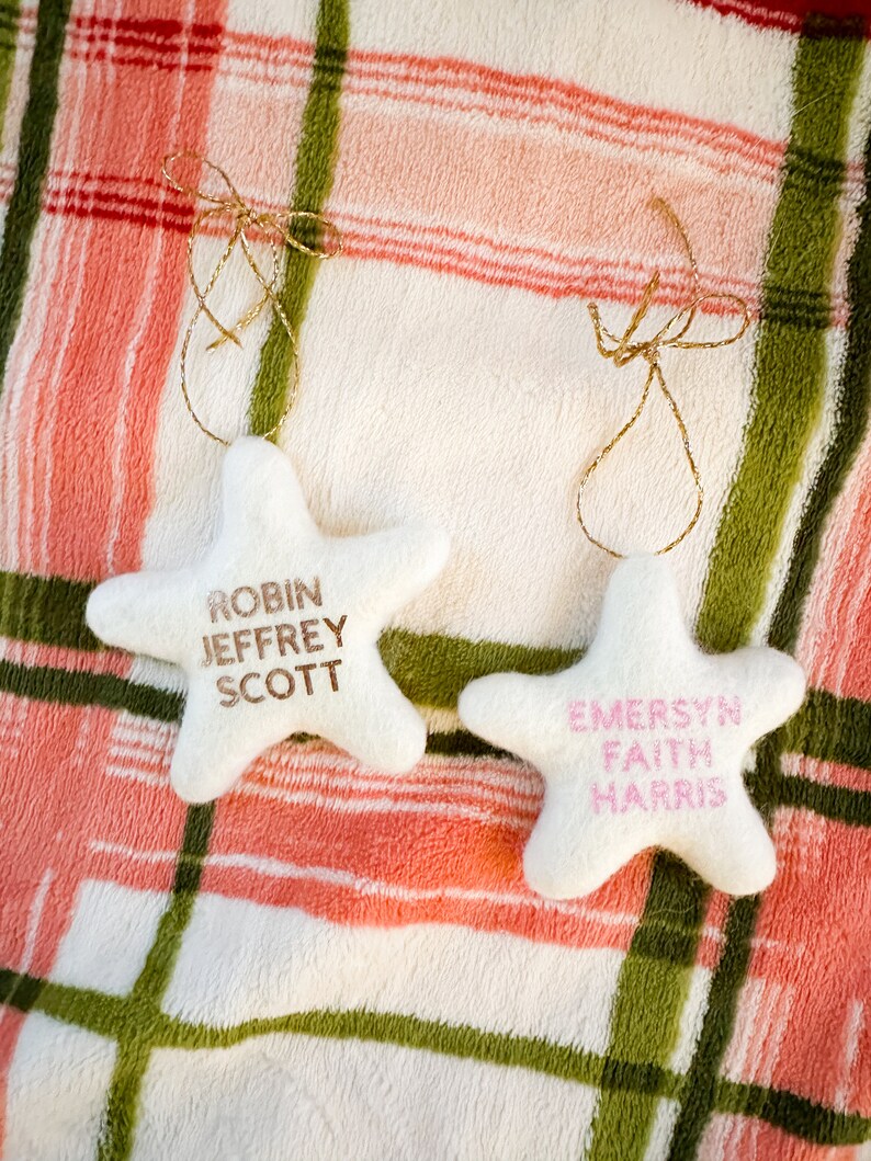 K&ouml;nnte beinhalten: Zwei wei&szlig;e sternf&ouml;rmige Ornamente mit goldener Schnur. Das Ornament links tr&auml;gt die Namen "Robin Jeffrey Scott" in goldenen Buchstaben. Das Ornament rechts tr&auml;gt die Namen "Emersyn Faith Harris" in rosa Buchstaben.