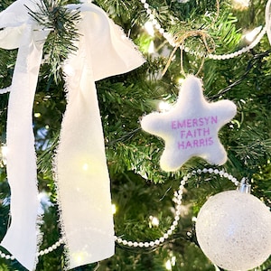 K&ouml;nnte beinhalten: Ein wei&szlig;er Filzstern-Anh&auml;nger mit pinkfarbener Aufschrift "Emersyn Faith Harris" h&auml;ngt an einem gr&uuml;nen Weihnachtsbaum mit wei&szlig;en Lichtern und einer wei&szlig;en Perlenkette.