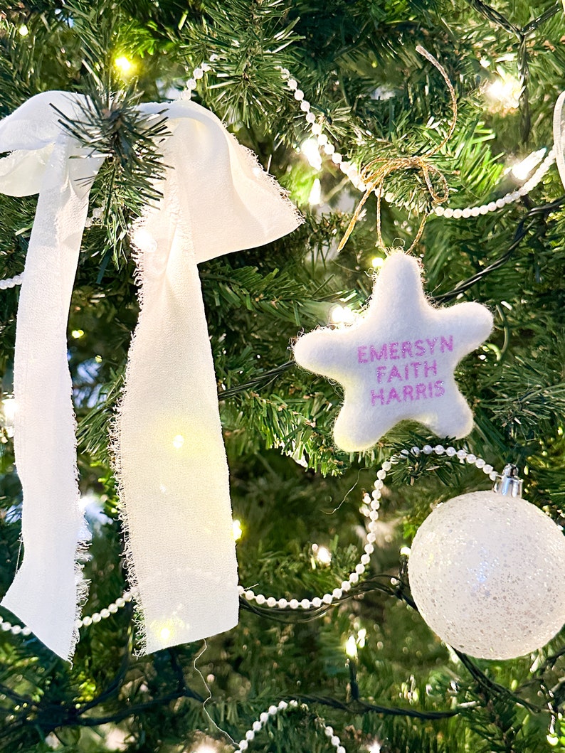 K&ouml;nnte beinhalten: Ein wei&szlig;er Filzstern-Anh&auml;nger mit pinkfarbener Aufschrift "Emersyn Faith Harris" h&auml;ngt an einem gr&uuml;nen Weihnachtsbaum mit wei&szlig;en Lichtern und einer wei&szlig;en Perlenkette.