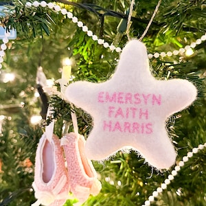 K&ouml;nnte beinhalten: Ein wei&szlig;er Filzstern-Anh&auml;nger mit rosa Stickerei, auf dem "EMERSYN FAITH HARRIS" steht, der an einem Weihnachtsbaum mit einer wei&szlig;en Perlenkette h&auml;ngt.