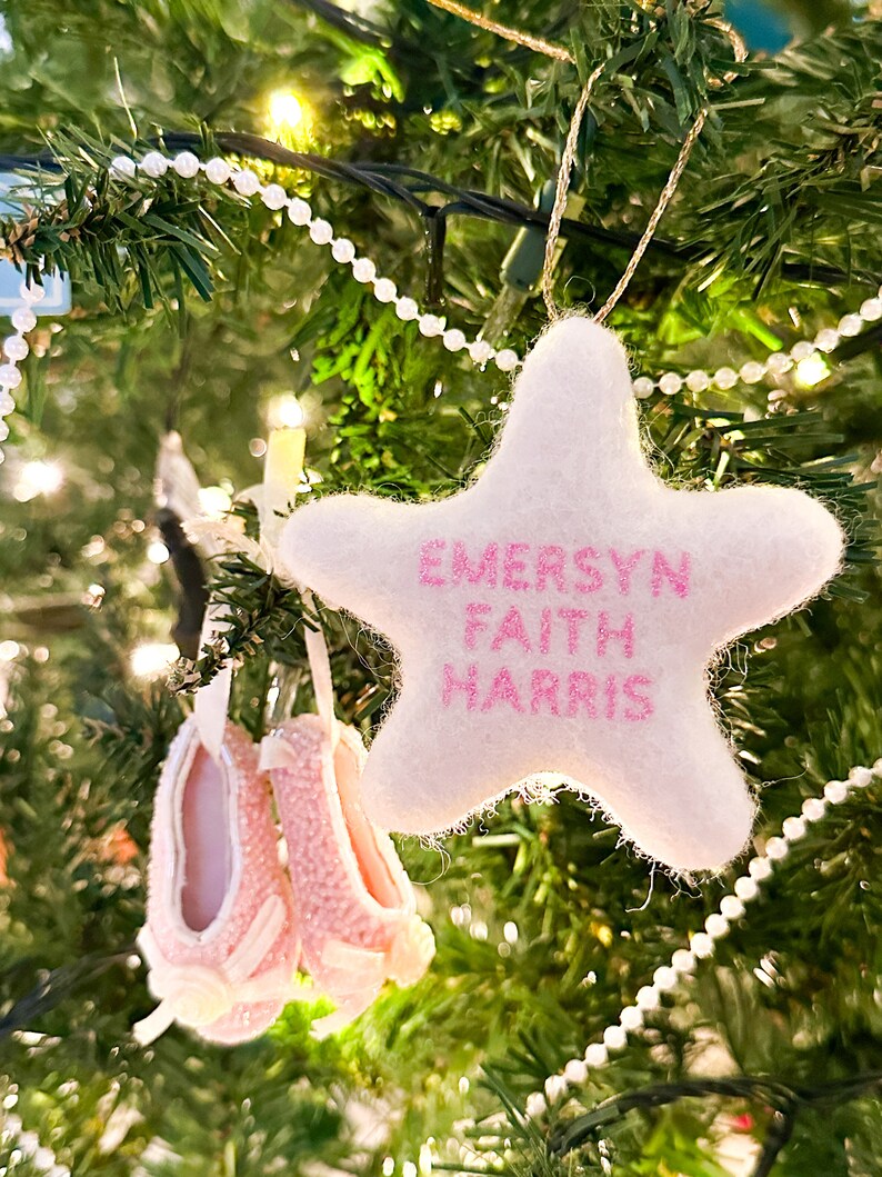 K&ouml;nnte beinhalten: Ein wei&szlig;er Filzstern-Anh&auml;nger mit rosa Stickerei, auf dem "EMERSYN FAITH HARRIS" steht, der an einem Weihnachtsbaum mit einer wei&szlig;en Perlenkette h&auml;ngt.
