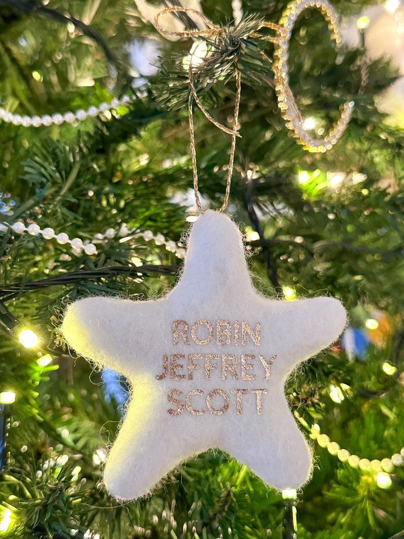 K&ouml;nnte beinhalten: Wei&szlig;er Filzstern-Anh&auml;nger mit goldenen Glitzerbuchstaben, auf dem "Robin Jeffrey Scott" steht, der an einem Weihnachtsbaum h&auml;ngt.