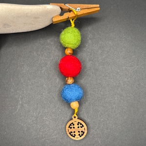 Op de afbeelding: Een houten wasknijper houdt een kleurrijk vilten balornament vast. Het ornament heeft een ontwerp van groene, rode en blauwe vilten ballen, met houten kralen en een kleine, ronde houten bedel met een kruisontwerp.