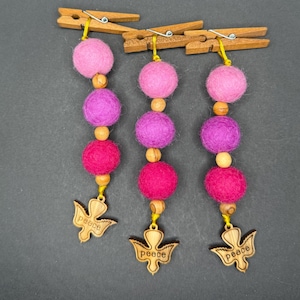 May include: Three decorative hanging ornaments with pink, purple, and fuchsia felt balls, wooden beads, and dove-shaped charms that read "peace". The ornaments are suspended from wooden clothespins.