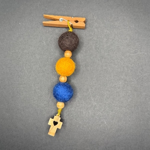 Może przedstawiać: Dekoracyjna klamerka do prania z sznurem filcowych kulek w kolorze brązowym, żółtym i niebieskim oraz drewnianym krzyżykiem. Klamerka do prania jest wykonana z drewna i ma metalową sprężynę. Krzyż ma wycięte serce.
