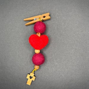 Op de afbeelding: Een decoratieve knijper met een reeks vilten ballen, een rood vilten hart en een houten kruisbedel. De vilten ballen zijn diep magenta van kleur en het hart is helderrood. Het kruis heeft een hartvormige uitsparing.