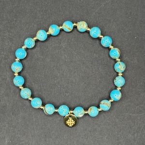 Puede incluir: Una pulsera de cuentas turquesas con detalles dorados y un dije dorado. La pulsera presenta cuentas redondas y moteadas en tonos turquesa y marrón. El dije dorado tiene un diseño de cruz.