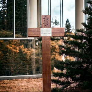 May include: A wooden cross with a silver plaque that reads "11.03.2006 - 01.10.2021" and several small heart cutouts. The cross is in front of a building with large windows and a pine tree.