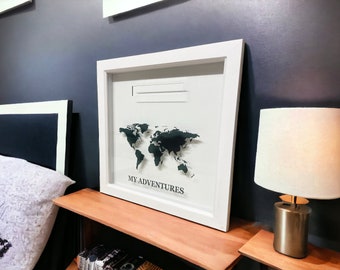 May include: White framed picture with a black world map and the text "MY ADVENTURES". The picture is hanging on a dark blue wall above a wooden shelf.