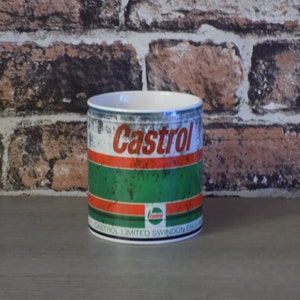 May include: A white ceramic mug with a distressed metal design featuring the Castrol logo in red and white. The mug has a green and red stripe design and the text "Castrol Limited Swindon England".