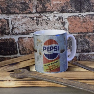 May include: White ceramic mug with a vintage Pepsi can design. The can is rusty and has the text "Pepsi Trademark" and "Let your taste decide. Take the Pepsi Challenge."