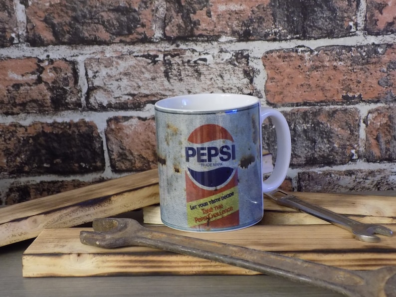 May include: White ceramic mug with a vintage Pepsi can design. The can is rusty and has the text "Pepsi Trademark" and "Let your taste decide. Take the Pepsi Challenge."