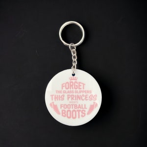 May include: White round keychain with a pink crown and text that reads "Forget the glass slippers this princess wears football boots".