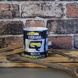 May include: A black ceramic mug with a yellow and white graphic of a vintage motor oil can. The can has the text "Duckhams Motor Oil" on it.