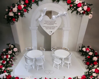 May include: A white wedding arch decorated with pink and red roses. The arch has a heart-shaped sign with the text "Our loved ones who are unable to be here in person today, We know you will look down on us on our Special Day, In memory of you we've saved you a seat, with the thought of you here makes our day complete."