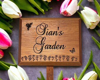 Può includere: Un cartello da giardino in legno con il testo "Sian's Garden" inciso su di esso. Il cartello è circondato da tulipani rosa e bianchi.