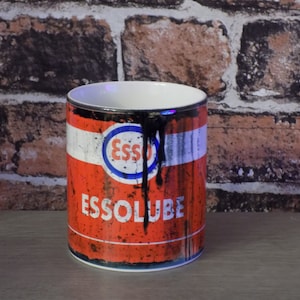 May include: A red and black ceramic mug with a vintage Esso oil can design. The mug has a white rim and handle. The design features the Esso logo and the text "Essolube".