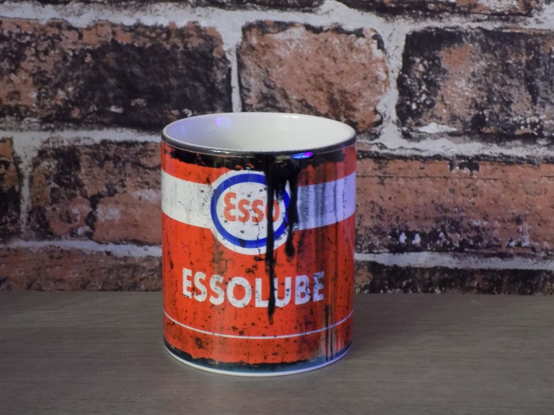 May include: A red and black ceramic mug with a vintage Esso oil can design. The mug has a white rim and handle. The design features the Esso logo and the text "Essolube".
