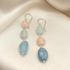 May include: A pair of silver dangle earrings with three gemstone beads. The top bead is pink, the middle bead is light blue, and the bottom bead is a larger, darker blue.