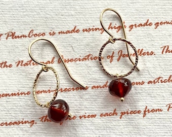 May include: A pair of gold earrings with a small, round, red gemstone dangling from each. The earrings have a delicate, textured design.
