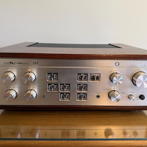 Könnte beinhalten: Ein Vintage-Vollverstärker Sansui 58A mit einer Frontplatte aus gebürstetem silbernem Metall, zahlreichen runden Drehknöpfen, Kippschaltern und einem Kopfhöreranschluss, untergebracht in einem dunklen Holzgehäuse mit schwarzer Lüftung.