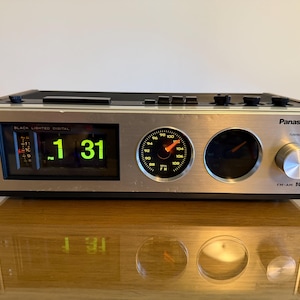 Può includere: Una radiosveglia vintage Panasonic FM-AM IC con pannello frontale argentato e inserti laterali effetto legno. Presenta un display digitale a palette che segna 1:31 PM, un quadrante analogico circolare per la radio FM con lancetta arancione.