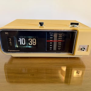 May include: A beige Panasonic flip clock radio with a black display panel showing the time 10:39 AM. The device features a horizontal radio frequency dial for AM and FM bands, two black control knobs on the top, and a band selector switch.