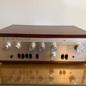 Op de afbeelding: Een Luxman solid-state geïntegreerde versterker met een geborsteld zilverkleurig metalen voorpaneel met meerdere ronde bedieningsknoppen, zwarte tuimelschakelaars en een hoofdtelefoonaansluiting, gevat in een donkerbruine houten behuizing.