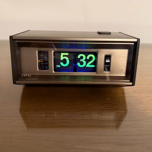 May include: A vintage Copal flip clock displaying 5:32 PM in bright green digits. The clock has a black and gold-toned metal casing. The clock is on a wooden surface.