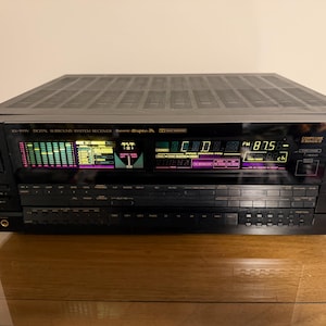 May include: A black JVC RX-999V digital surround sound system receiver. The receiver has a display with green, yellow, and purple lights, and a digital tuner showing 87.5 MHz. It has multiple buttons and controls.