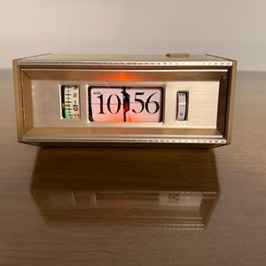 May include: A vintage flip clock with a gold-tone metal frame. The clock displays the time as 10:56 AM with white numbers on a black background. The clock has a small green and white indicator on the left side and a small dial on the right.