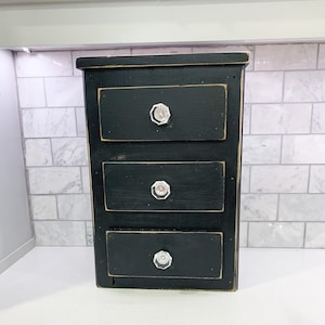 May include: A small, black, three-drawer cabinet with crystal knobs. The cabinet has a distressed finish, giving it a rustic or vintage appearance. The drawers are rectangular and evenly spaced. The cabinet is likely made of wood.