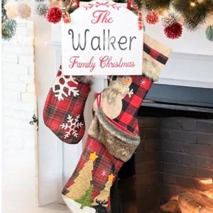 May include: A Christmas stocking holder with three plaid stockings. The stockings have snowflake, snowman, and Christmas tree designs. A sign above the stockings reads "The Walker Family Christmas". The holder is green and the sign is white.