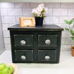 May include: A small, black, four-drawer cabinet with crystal knobs. A small wooden framed picture and a potted plant with white flowers sit on top. A small lemon tree is to the right.