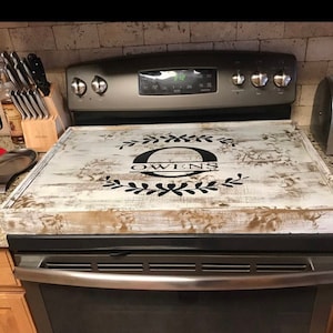 May include: A distressed white wooden stove cover with the name "OWENS" in black script. The cover features a decorative floral design and is designed to fit over a standard stove top. The wood has a rustic, weathered appearance.