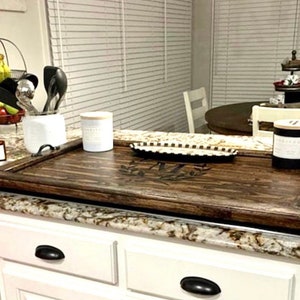 May include: A dark brown wooden stovetop cover with a decorative design. The cover is placed on a white countertop with a granite pattern. There are three white ceramic containers and a black candle on the countertop.