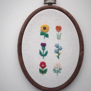May include: Embroidered hoop art featuring six different flowers in various colors, including a yellow sunflower, a red tulip, a purple thistle, a blue forget-me-not, a red poppy, and a white daisy.