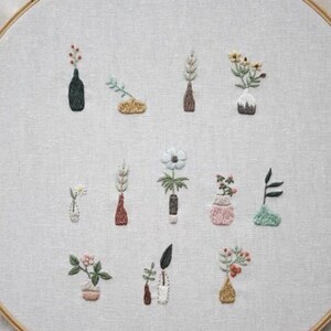 Puede incluir: Un aro de bordado blanco con 12 diseños florales bordados diferentes. Los diseños son todos de diferentes colores y estilos, y están todos dispuestos en un patrón circular. El aro está hecho de madera y tiene un acabado natural.