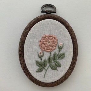 May include: A brown wooden embroidery hoop with white linen fabric inside. The fabric features a hand-stitched pink flower with green leaves.