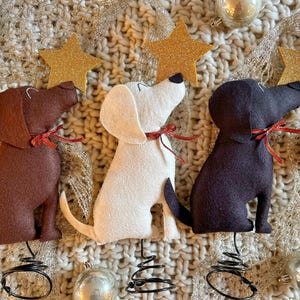 May include: Three felt dog ornaments in brown, white, and black, each with a red ribbon collar and a gold glitter star. The dogs sit on a knitted surface with silver tinsel and glass ball ornaments, perfect for holiday decor.
