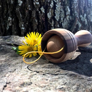 May include: A wooden toy with a ball on a string. The toy is made from different types of wood, including walnut and maple. The ball is made of maple. The toy is lying on a rock with yellow flowers.