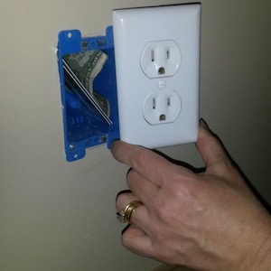 May include: A white electrical outlet cover is held against a blue electrical box in a wall. A hand with rings is holding the cover. A dollar bill is visible inside the electrical box.