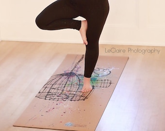 May include: A woman wearing a brown tank top and black leggings performs a yoga pose on a cork yoga mat with a painted design of a birdcage. The mat has the text "STRAW" printed on it. The text "LeClaire Photography" is printed on the floor.