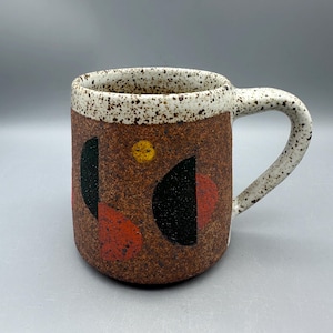 May include: A ceramic mug with a speckled white rim and handle, featuring a textured brown body decorated with black semi-circles, red shapes, and a small yellow circle. The mug has a cylindrical shape and a sturdy side handle.