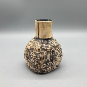 May include: A small, round ceramic vase with a textured, woven pattern in black and white. The vase has a narrow neck and a wide base.